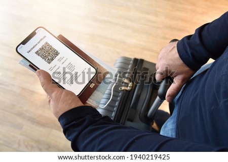 Travel and tourism concept during coronavirus pandemic. Man holds smartphone with digital certificate of vaccination against Covid-19, passport, face mask in one hand, and a travel bag in the other.