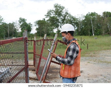An Indonesian engineer wearing hard hat helmet checking oil derrick field. An Indonesian engineer in work and white helmet checking oil pumping unit at oil field, using tablet.