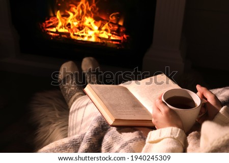 Woman with book and cup near fireplace indoors, closeup. Cozy atmosphere