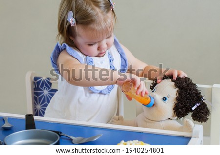 Funny blonde toddler girl feeding a doll at home, taking care of a doll, pretend play babysitting. Kitchen toys simulation, educational and role play toys for young kid  Royalty-Free Stock Photo #1940254801