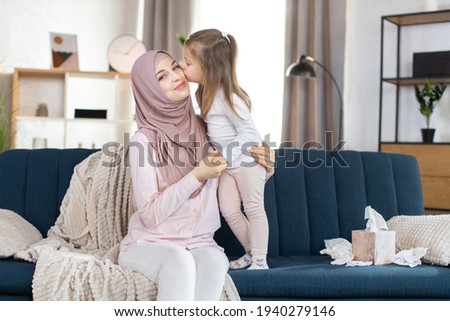 Muslim family mom and child. Cute little girl, kissing her smilin mom in hijab in cheek with love. Happy young Arabian mother having fun with her child girl, spending free time at home.