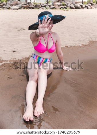 A lady in a large sun hat and bikini declines to have her photo taken at the beach, preferring to go incognito and not have evidence on social media.