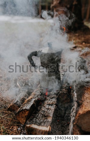 Preparing coffee in old vintage coffee can in bonfire in camping adventure. Early spring vacations. Blurred brown grass and green forest in background Coffee can disappearing in white smoke. Close-up