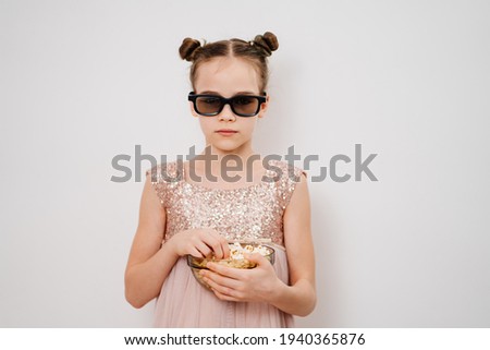 A teen girl with glasses to watch sad 3D movies eats popcorn stands against a white wall. Home or online cinema. harmful snacking.