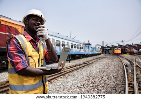 Portrait of handsome Africa American engineering using walkie talkie for control labor with laptop in front of train garage.  Back view of contractor on background of outdoor old train garage.