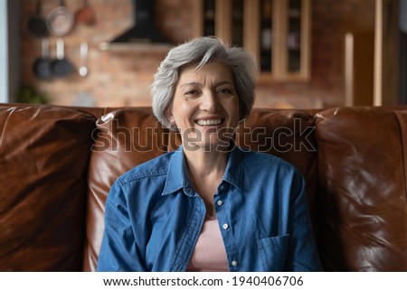 Active old age. Portrait of optimistic retired hispanic female clothed in stylish casual demonstrating perfect white teeth in beautiful smile. Energetic senior latin woman sit on couch look at camera