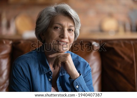 Smart look. Profile picture of attractive old latin lady of retirement age enjoy self beauty care look at camera with smile. Beautiful elegant elderly female posing for portrait on cozy sofa at home