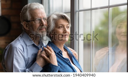 Looking in future. Loving senior man husband embrace aged wife from behind support encourage share feeling of beloved woman. Elderly spouses hug look at window meet twilight years together. Copy space Royalty-Free Stock Photo #1940409940