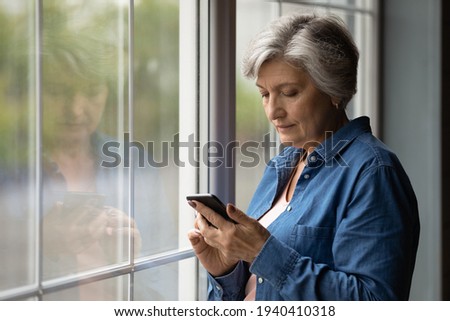 Important message. Serious aged latin female pensioner stand by picture window at home hold cell text in messenger app. Focused older woman thinking on email received from bank write answer. Copyspace Royalty-Free Stock Photo #1940410318