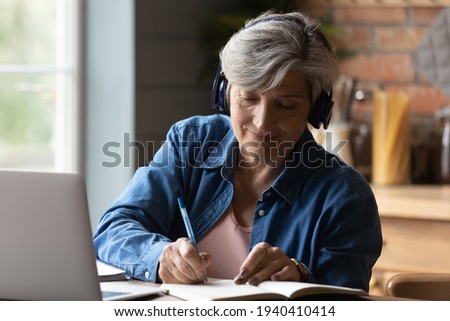 Diligent aged student. Focused elderly hispanic woman in headphones watch webinar on laptop screen take notes to paper notebook. Mature old lady in headset study online take part at education training Royalty-Free Stock Photo #1940410414