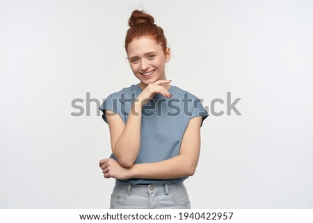 Nice looking woman, beautiful redhead girl with hair bun. Wearing blue t-shirt. Touching her chin, lays her head on a hand. Broadly smiling. Watching at the camera, isolated over white background