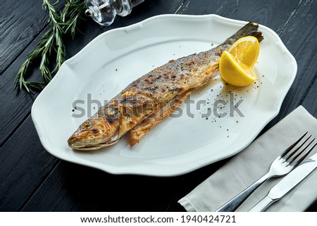 Whole grilled trout on charcoal fire, served in a white plate with lemon on a dark wood background.