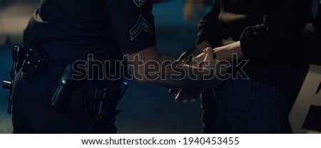 CLOSE UP Police officer handcuffs a suspect near police car, African-American Black criminal. Lights flashing in the background. Shot with anamorphic lens