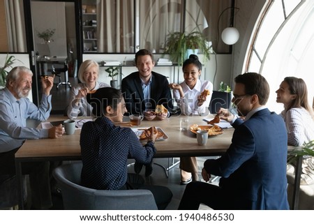 Smiling diverse employees sit in office have fun enjoy pizza on work lunch break together. Happy multiethnic colleagues coworkers laugh joke eating Italian fast food meal, drink coffee at workplace. Royalty-Free Stock Photo #1940486530