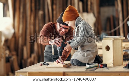 Professional craftsman teaching little son to use drill while making together wooden bird house in carpentry workshop