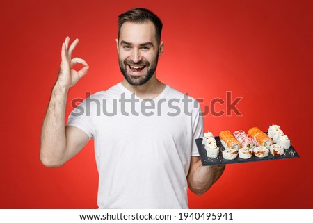 Excited young bearded man 20s wearing casual white t-shirt showing ok okay gesture hold makizushi sushi roll served on black plate traditional japanese food isolated on red background studio portrait