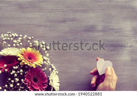 Orange and burgundy gerberas and white baby breath flowers on grey wood background with copy space, place for your text. Flower arrangement, floral corner composition. Hand hold paper heart.