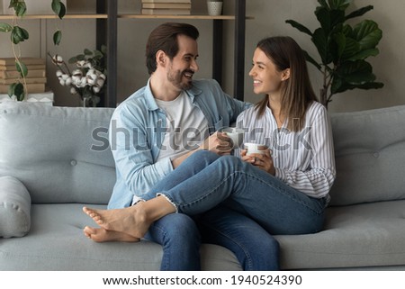Happy young couple relaxing on cozy couch at home, holding cups, drinking hot beverages, tea or coffee together, laughing man and woman enjoying leisure time, chatting, sharing news, having fun Royalty-Free Stock Photo #1940524390