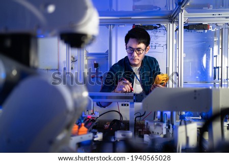 Young Asian male electrical engineer in glasses using a digital multimeter in hand checking voltage to fix an industrial machine with a blurred of automation robotic arm machine in the foreground. Royalty-Free Stock Photo #1940565028