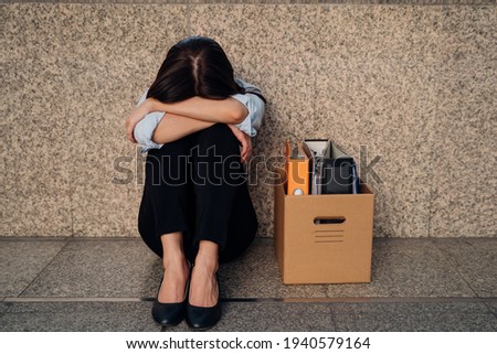 Unrecognized young woman hiding her face after being laid off from job due to economic recession sitting with carton of belongings on floor and crying Royalty-Free Stock Photo #1940579164