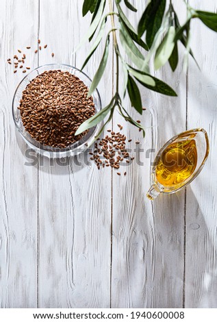 Linseed oil and heap of flax seeds on a white wooden table. Modern still life glass bowl with oil, linseed and olive branch, copy space.