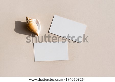 Still life scene with white shells on beige background in sunlight and blank business, greeting card, invitation mockup. Long harsh shadows. Flat lay, top view.
