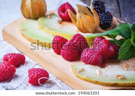 Mix of raspberries, blackberries, mint, physalis and melon on wooden cutting board over wooden table.