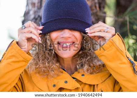 Cheerful adult beautiful woman portrait covering his eyes with winter cup - happy people enjoy outdoor leisure activity - pretty smile and teeth concept - middle age caucasian lady in the woods 