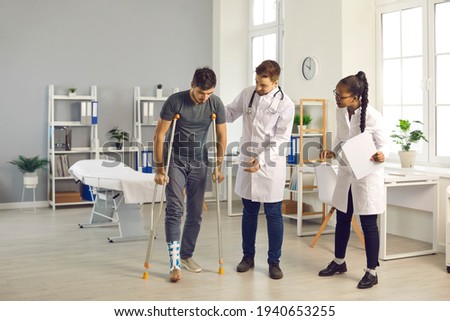 Team of doctors of different nationalities in the hospital room helps their patient with a broken ankle, who walks on the cauldrons. Concept of treatment of serious physical injuries. Royalty-Free Stock Photo #1940653255