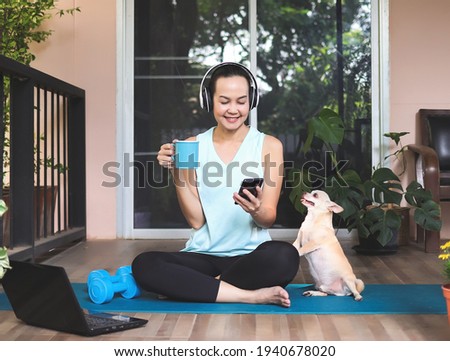Portrait of Asian woman wearing headphones, holding coffee cup, using smartphone, sitting  on yoga mat in balcony  with computer laptop dumbbell and Chihuahua dog. Yoga exercise training online