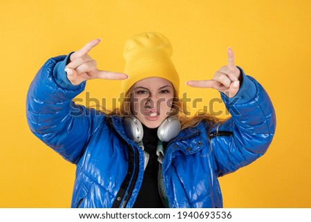 An emotional Caucasian woman makes the rock and roll sign, says I'm going to rock this party, screams loud, wears round spectacles, casual to pants, stands on yellow background