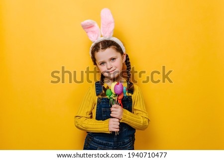 Beautiful smiling preschool girl kid with bunny fluffy ears, holds painted eggs on sticks, happy look at camera, going to celebrate Easter, isolated on yellow studio background. Spring holiday concept