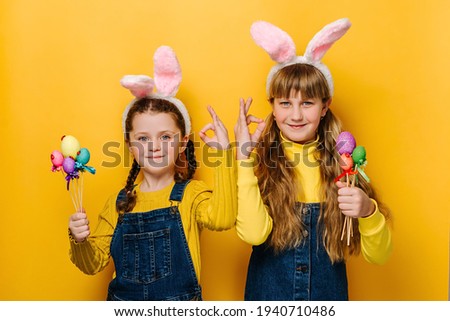 Portrait of cute beautiful little sisters girls in pink bunny fluffy ears holding small Easter colored eggs, makes okay gesture, says ok, isolated on yellow studio background. Spring holiday concept