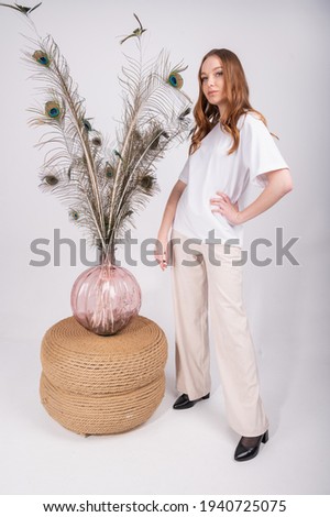 Portrait of young beautiful woman with long hairstyle, wearing white t-shirt and light pants over peacock feather background. High quality photo
