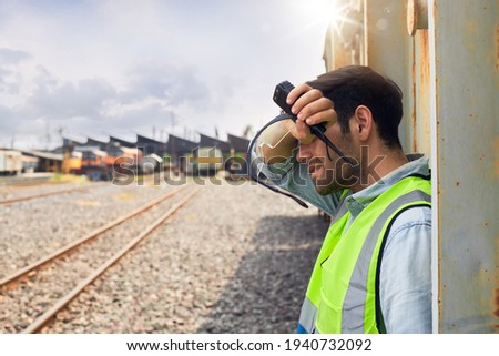 A Railway engineer or Rail transport technician wearing a green safety vest is standing to rest or working outdoors beside a freight train on a hot and sunny day. Royalty-Free Stock Photo #1940732092