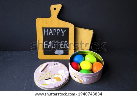 Against a black background, there is a festive metal box with colorful Easter eggs next to a wooden note board and a paper notepad in the background.  Photo view from the top.