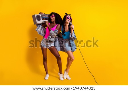 Photo of two pretty girlfriends wear denim clothes dark glasses holding boombox singing empty space isolated yellow color background
