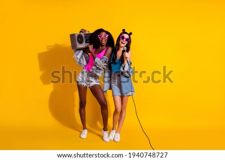 Full body photo of two happy women sing hold boombox summer mood wear jeans jacket shorts isolated on yellow color background