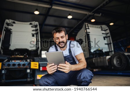 Smiling tattooed bearded blue collar worker in overalls using tablet to check on delivery while crouching in garage of import and export firm. In background are trucks. Royalty-Free Stock Photo #1940766544