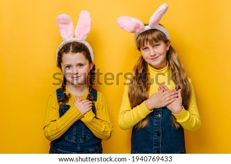 Portrait of two happy smiling girls kid in pink bunny fluffy ears put folded palms on heart, isolated over yellow studio background. Concept easter and sign of sincere kind person, feeling thankful