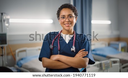 Hospital Ward: Portrait of Posing Beautiful Black Female Head Nurse, Doctor, Surgeon Smiles Charmingly and Kindly Looks at Camera. Modern Clinic with Advanced Equipment and Professional Staff