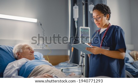 Hospital Ward: Friendly Head Nurse Talks with Elderly Patient Resting in Bed. Physician Uses Tablet Computer, Does Checkup, Ask Health Care Questions. Old Man Fully Recovering after Successful Surgery Royalty-Free Stock Photo #1940771293