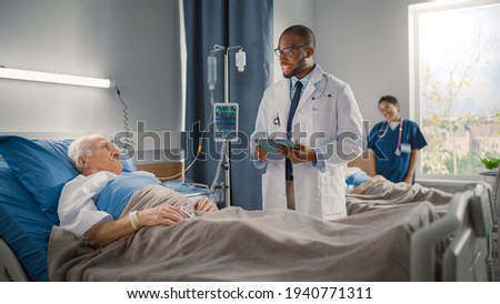 Hospital Ward: Friendly Black Doctor Talks with Elderly Caucasian Patient Resting in Bed, Asks Health Care Questions. Doctor Uses Tablet Computer. Old Man Fully Recovering after Successful Surgery Royalty-Free Stock Photo #1940771311
