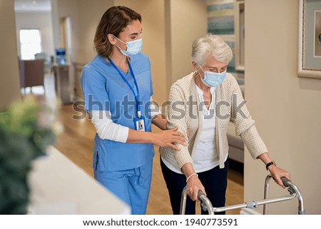 Smiling nurse with face mask helping senior woman to walk around the nursing home with walker. Young lovely nurse helping old woman with surgical mask for safety against covid-19 using a walking frame Royalty-Free Stock Photo #1940773591