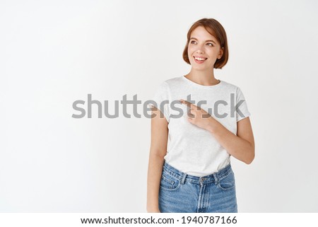 Beautiful natural girl without make-up, white teeth, smiling and pointing left at logo, showing banner, standing in t-shirt on studio background