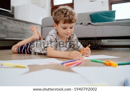 A school-age boy lies on the living room floor and paints pictures on white sheets of paper with colored markers. The boy is spending his afternoon free time at home.