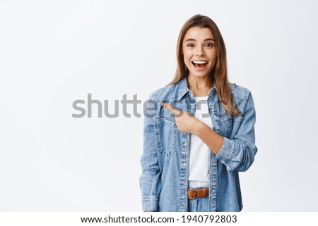 Wow this is good, look here. Smiling cute girl with blond hair pointing finger left at copy space and staring excited at camera, showing awesome promo offer, white background Royalty-Free Stock Photo #1940792803