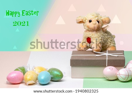 Easter greeting card. Close-up of colorful eggs, a gift box with a easter lamb on it on a multicolour table in front of abstract pastel background with inscription Happy easter 2021. Copy space. Relig