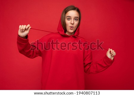 Photo shot of beautiful surprised happy smiling brunette female teenager wearing trendy red hoodie standing isolated over red background wall looking at camera