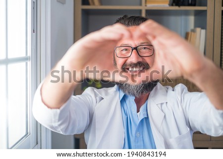 Close up portrait of smiling young caucasian male nurse or GP in white medical uniform show heart love hand gesture. Happy man doctor show support and care to patients or client in hospital.